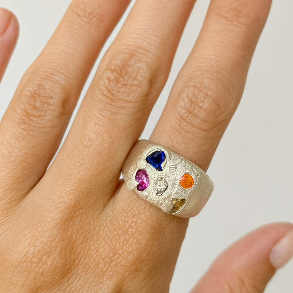 Hand wearing a ring with colorful gemstones on a neutral background
