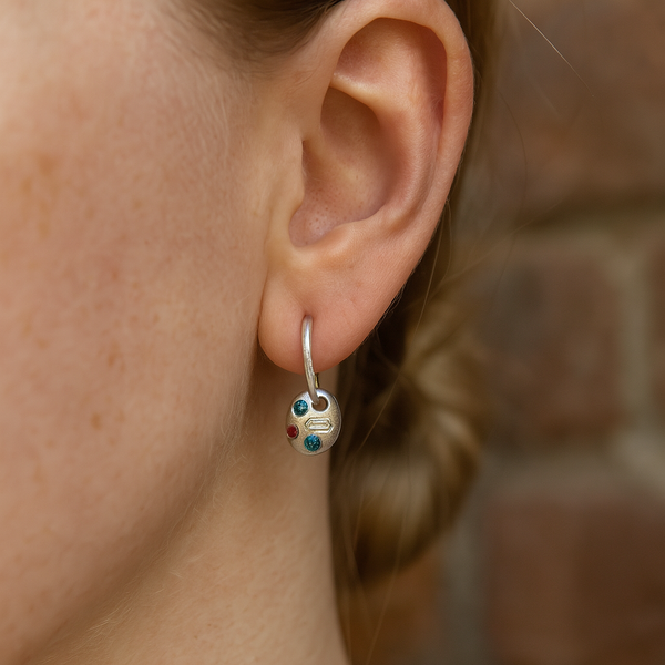 Close-up of an ear wearing a small hoop earring with colored stones against a blurred brick wall background.