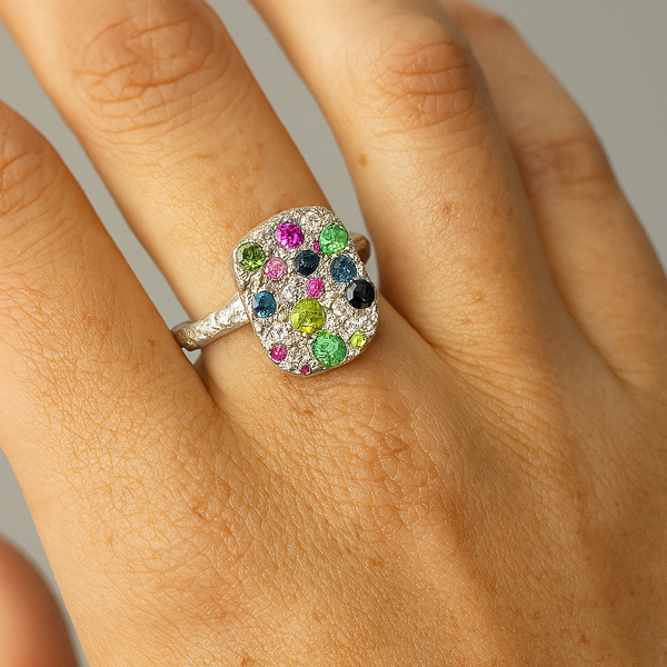 Close-up of a hand wearing a colorful gemstone ring on a neutral background